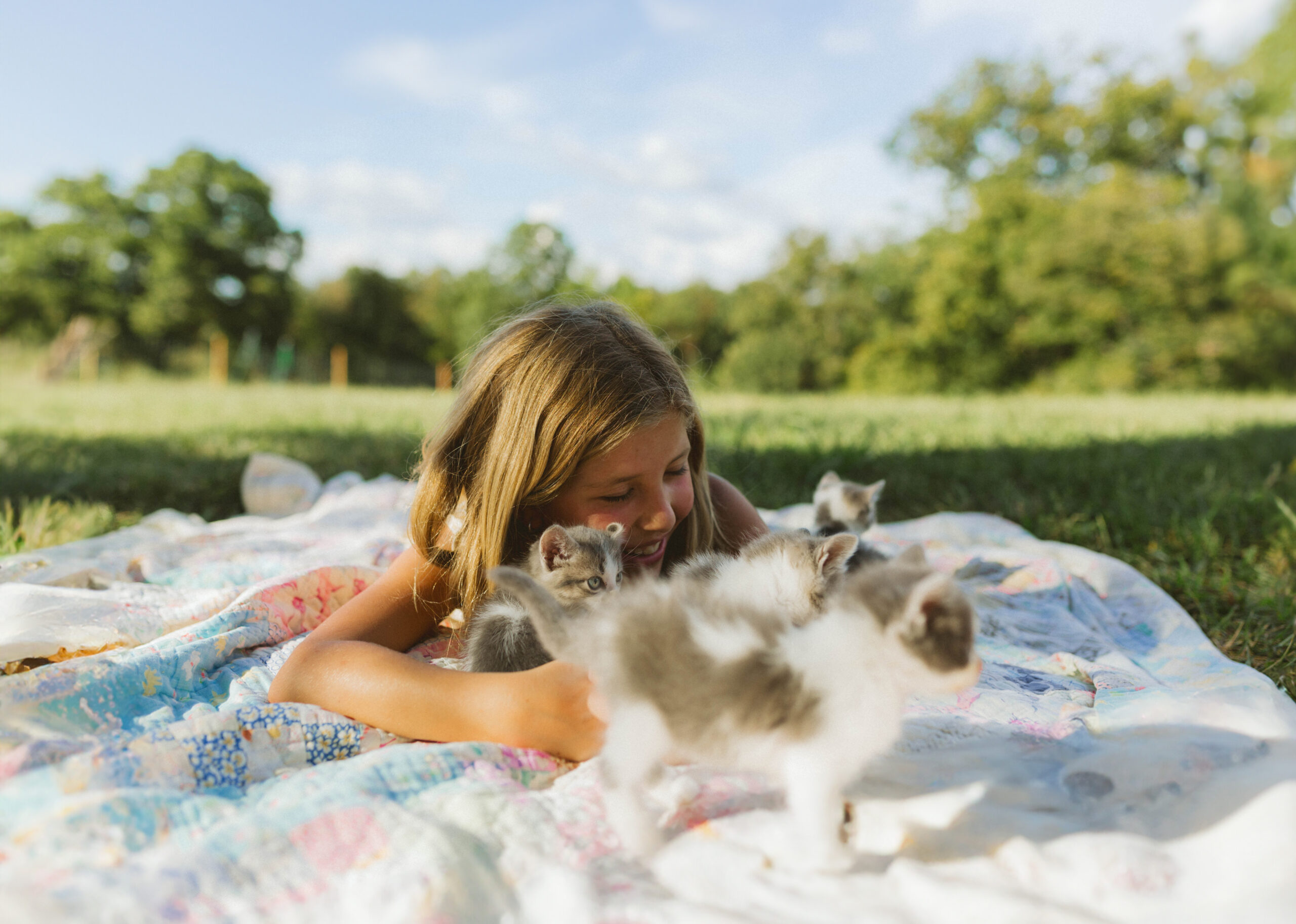 8-year-old girl laying on a blanket playing with kittens outdoors, smiling and enjoying a playful moment.