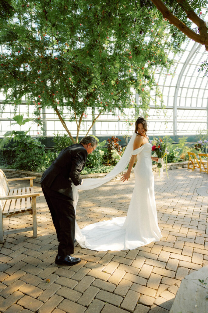 bride smiles back at her dad as he fixes her train before walking down the aisle at garfield park conservatory in chicago