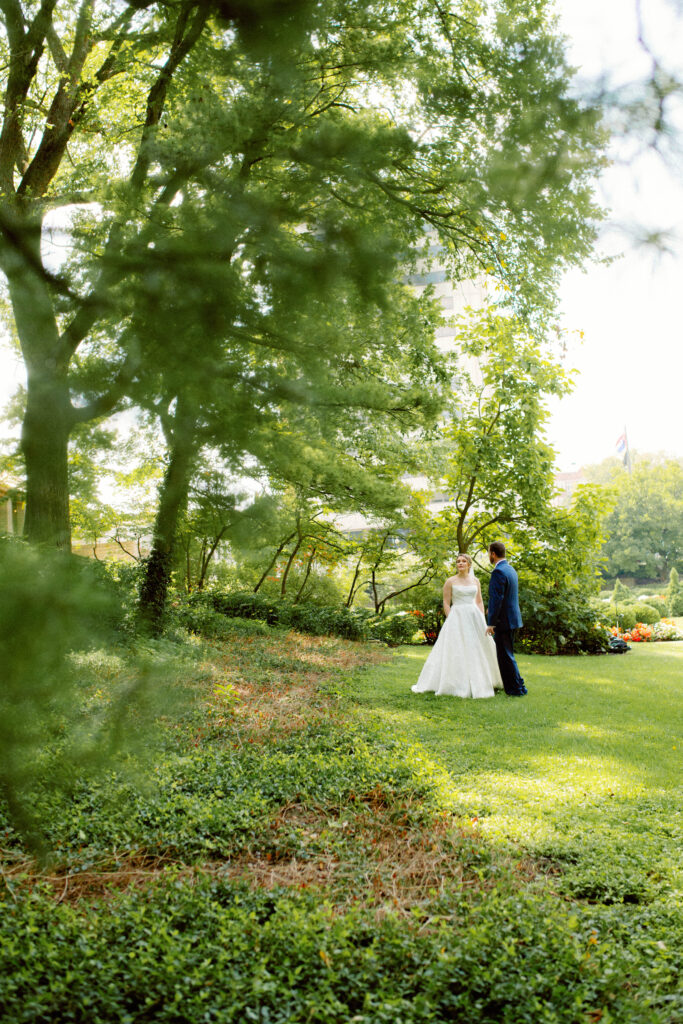 bride looks towards scenery as groom looks at her in a natural moment after their first look at carnahan memorial garden in jefferson city missouri