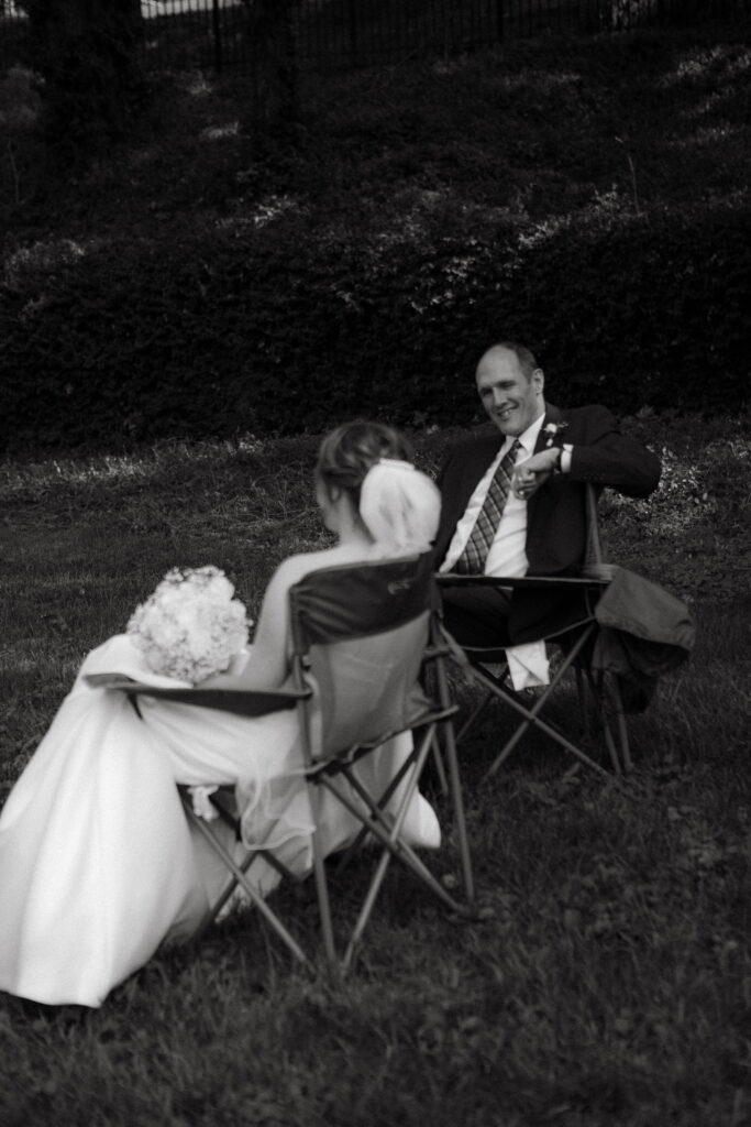 bride and her dad sit in lawn chairs as dad smiles big at her as they wait for the ceremony at carnahan memorial garden in jefferson city missouri